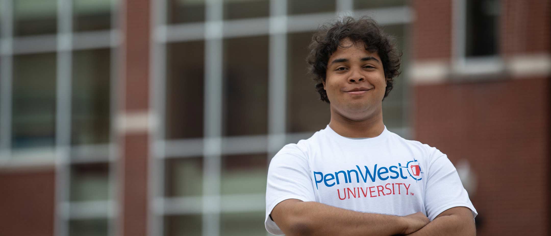 A student with folded arms smiling in front of campus building.